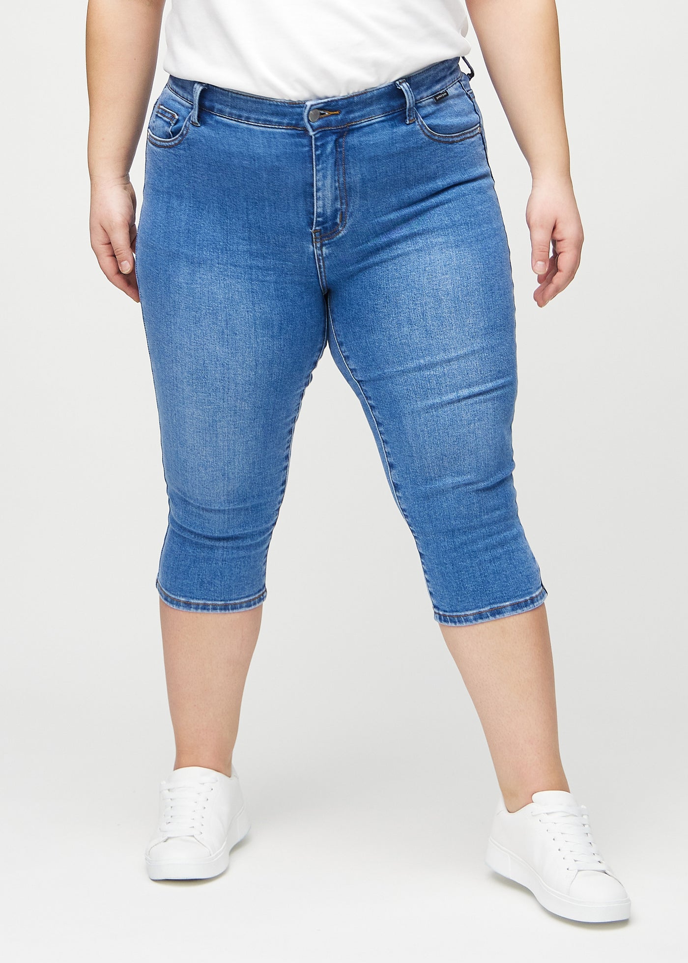 Denim skinny capris, modelnavn Rivers, som sidder tæt til benet og slutter lige under knæet på en plus-size model, set forfra.